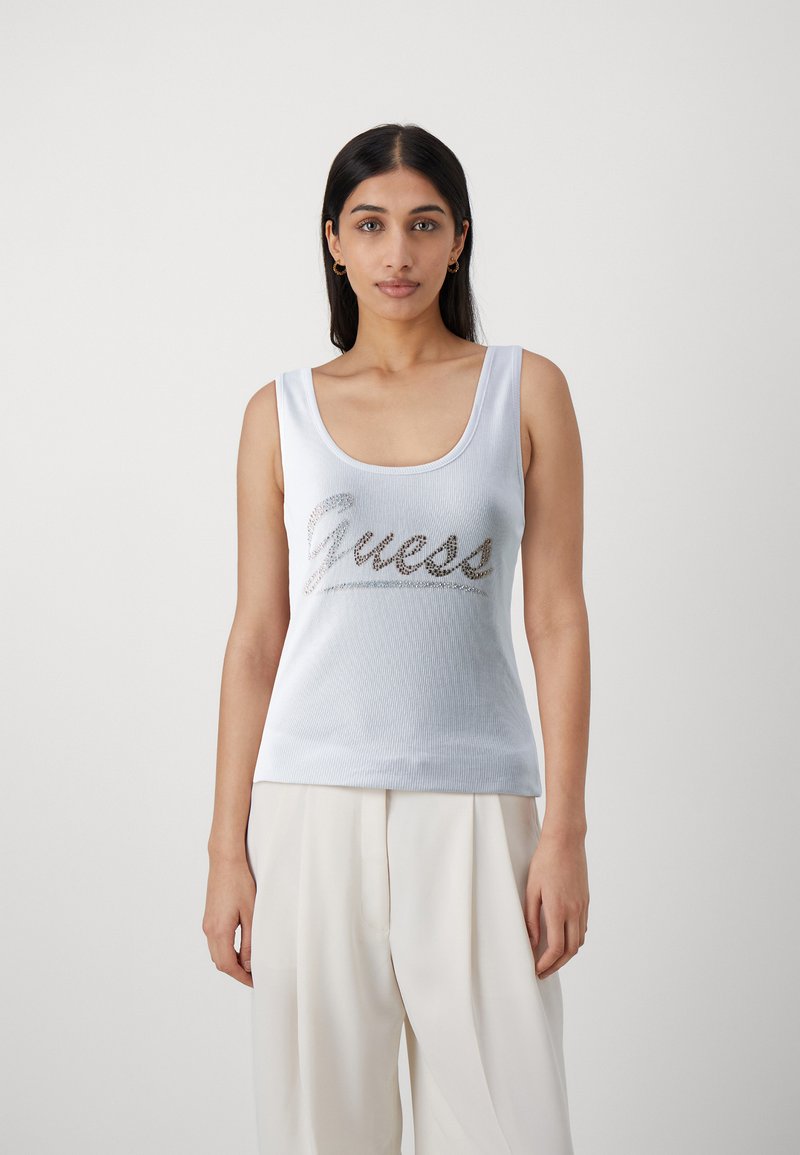 Camiseta sin mangas blanca con textura acanalada que presenta un logo metálico plateado "Guess" en el pecho; escote redondeado y tirantes anchos.