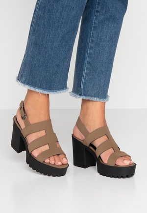 Brune højhælede sandaler med tre brede remme, justerbar ankelspænde, platformsål og tekstureret sort bund. Filtrerede blå cowboybukser.