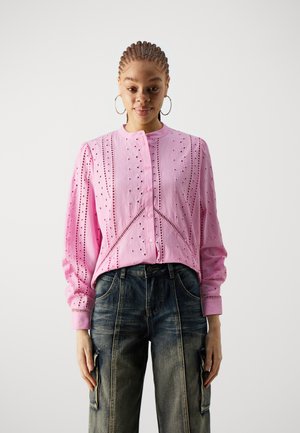 Pink blouse with geometric cutouts, long sleeves, and a round collar, paired with dark denim pants featuring side pockets.