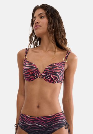 Frau trägt einen rosa, schwarzen und lila Bikini mit Tigerstreifen und goldenen Ringdetails an den Schulterträgern vor einem einfarbigen Hintergrund.
