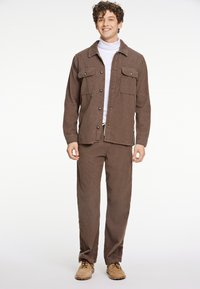 Camisa chaqueta de corduroy marrón con dos bolsillos en el pecho, combinada con pantalones marrones a juego y zapatos beige, con un cuello alto blanco debajo.