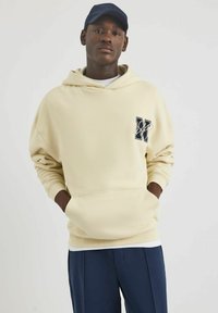 Kremowa bluza z kapturem z kieszenią z przodu i akcentem w formie logo. Charakteryzuje się prążkowanymi mankietami i kapturem. W połączeniu z granatowymi spodniami. Gładka tekstura, casualowy fason.