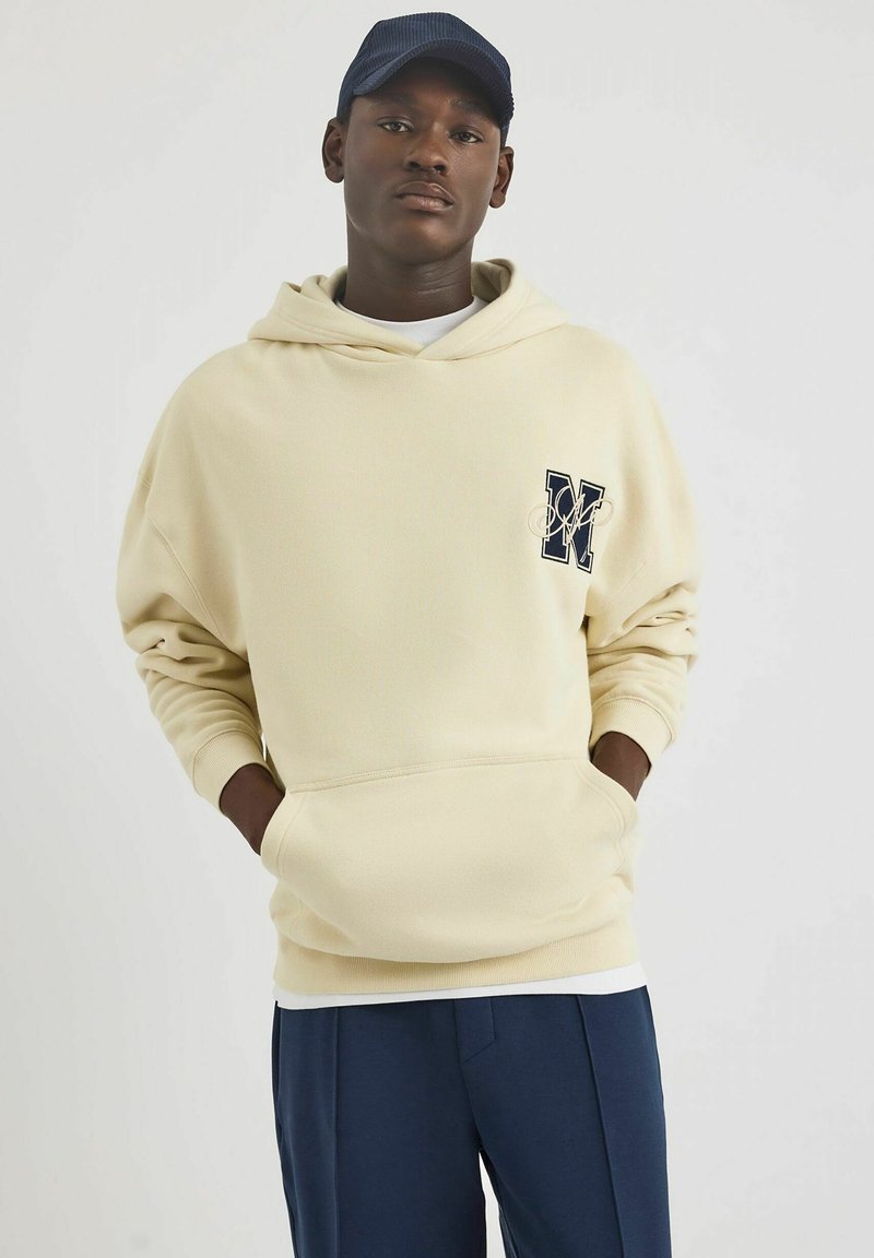 Kremowa bluza z kapturem z kieszenią z przodu i akcentem w formie logo. Charakteryzuje się prążkowanymi mankietami i kapturem. W połączeniu z granatowymi spodniami. Gładka tekstura, casualowy fason.