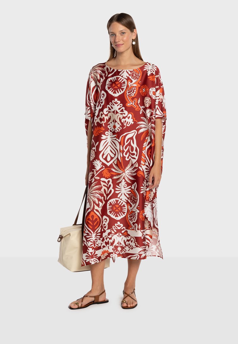 Robe à motifs rouges et blancs avec manches larges, coupe au genou et motif floral. Le modèle tient un sac crème et porte des sandales marron.
