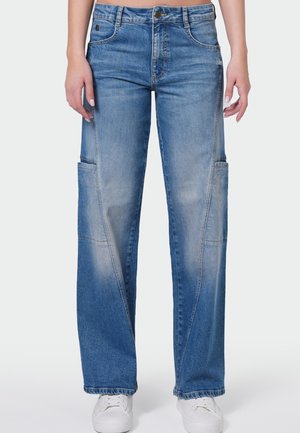 Blauwe rechte denimspijkerbroek met zijzakken, gedragen met witte sneakers door een persoon die voor een effen achtergrond staat.