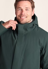 Dark green waterproof jacket with a hood, smooth texture, featuring a high collar and zipper closure. Accented with adjustable drawstrings.