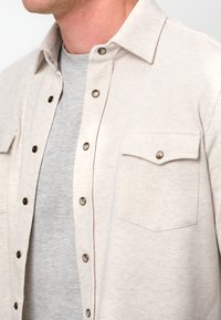 Camicia beige con colletto, dotata di bottoni scuri e due tasche sul petto, indossata sopra una maglietta grigia a girocollo. Texture morbida.
