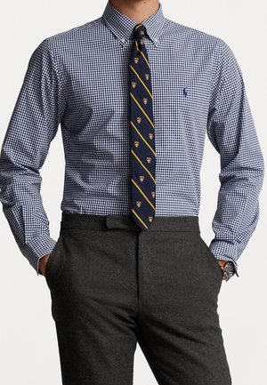 Homme portant une chemise à carreaux bleue et blanche, une cravate rayée marine avec des emblèmes de bouclier, un pantalon gris foncé et une montre-bracelet, les mains dans les poches.