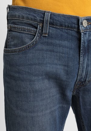 Nahaufnahme des Taillen- und oberen Oberschenkelbereichs blauer Lee-Jeans, getragen mit einem senfgelben Oberteil, zeigt die vorderen Taschen und die Details des Jeansknopfs.
