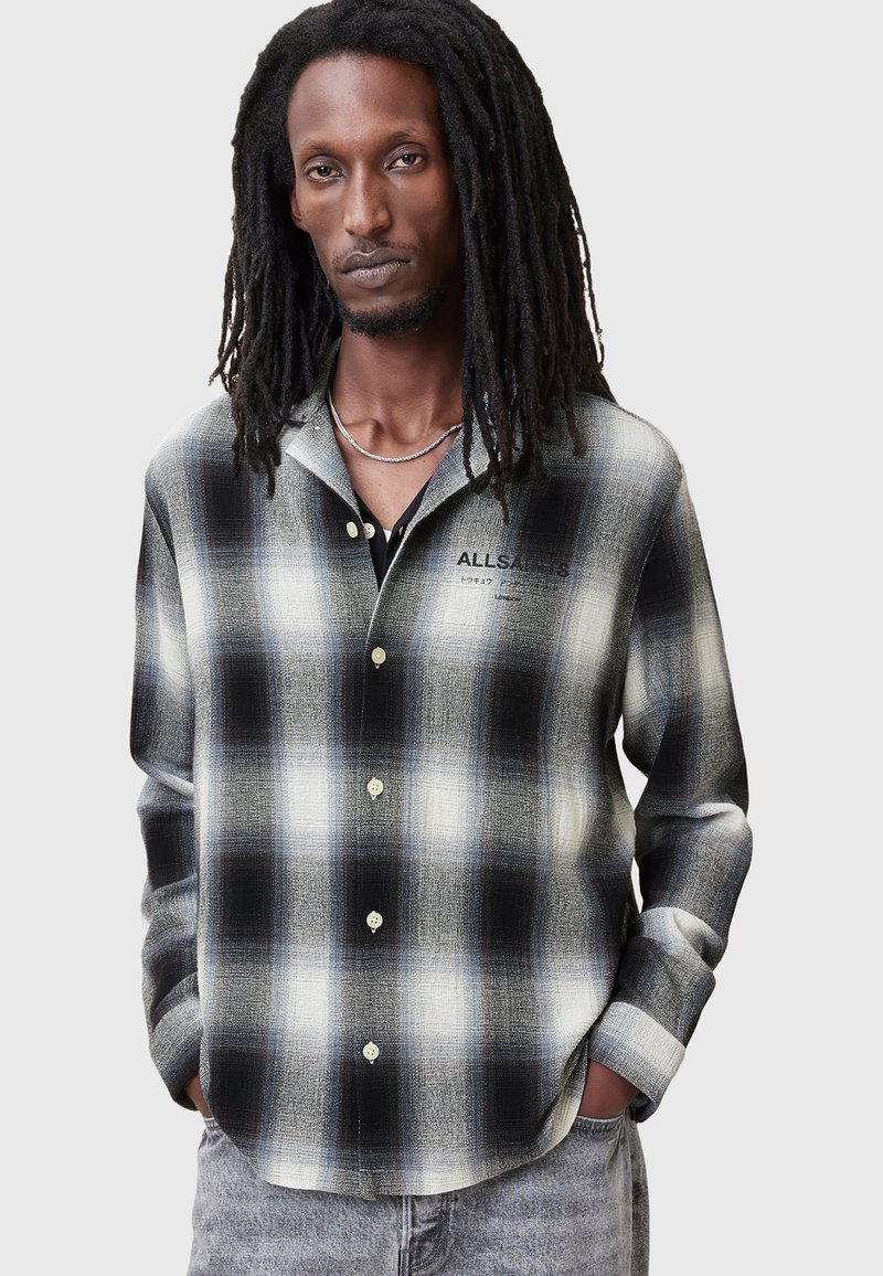 Camisa de cuadros en blanco y negro con botones en la parte delantera, tela de textura suave, mangas largas y corte relajado. Logo impreso en el pecho.