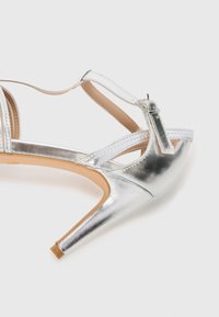 Silberne High-Heel-Sandale mit einem eleganten metallischen Finish, Kreuzriemen-Design, verstellbarem Knöchelriemen und einer beigen Sohle.