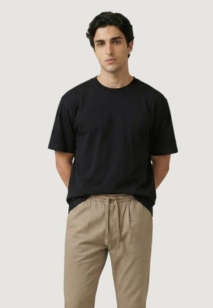 Uomo con capelli ricci neri, che indossa una maglietta nera a maniche corte e pantaloni beige con coulisse, su uno sfondo chiaro uniforme.