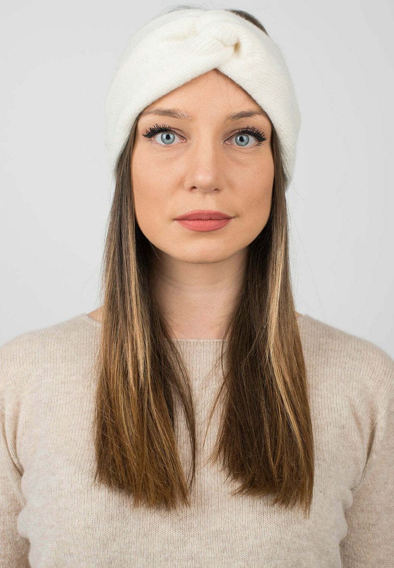 Fascia bianca lavorata a maglia con un design intrecciato, realizzata in materiale morbido. Indossata da una persona con capelli castani lisci, caratterizzata da una trama semplice.