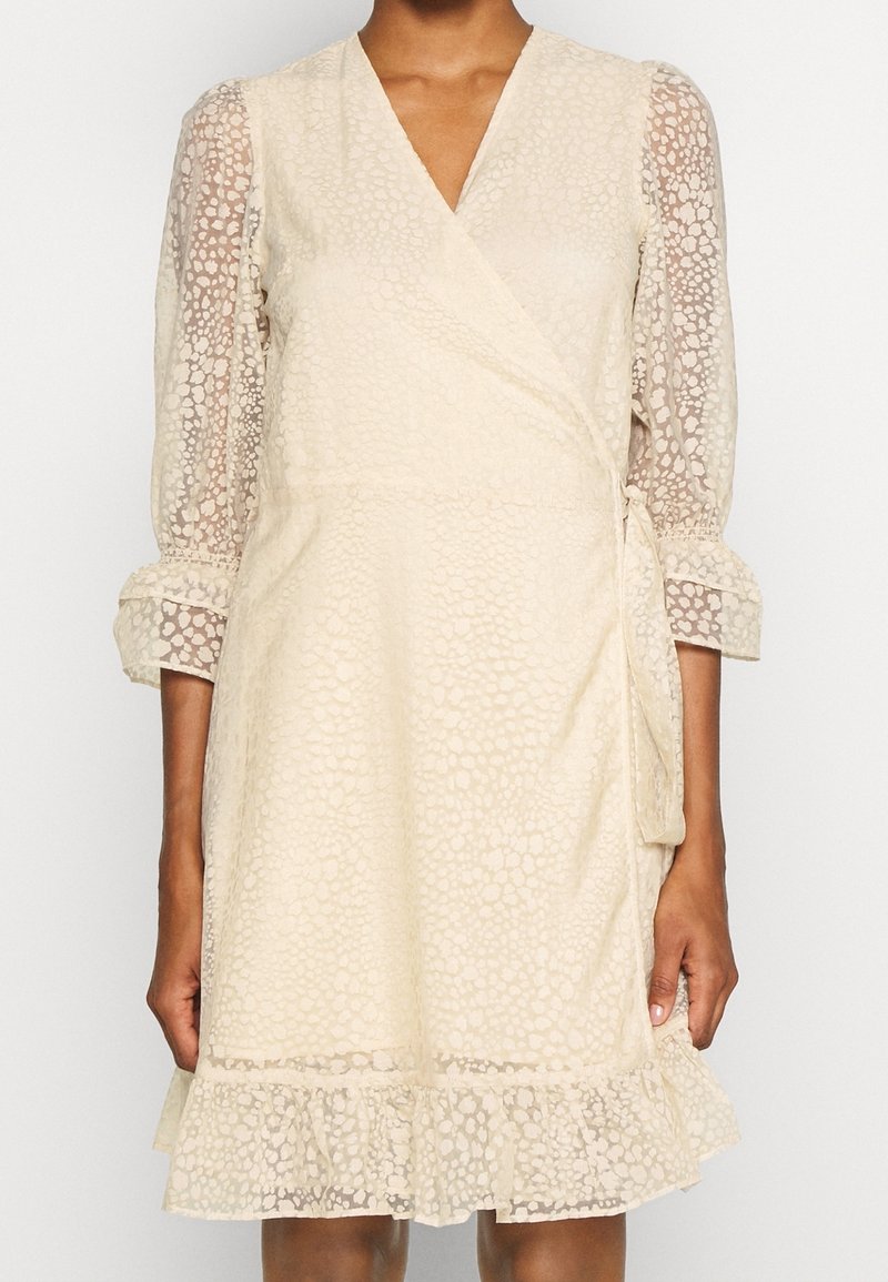 Robe enveloppante couleur crème avec des manches transparentes à motifs, un décolleté en V et un ourlet à volants. Présente des détails texturés ton sur ton tout au long.