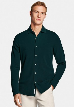 Mann trägt ein dunkelgrünes Hemd mit langen Ärmeln und hellbeige Hosen, steht mit einer Hand in der Tasche vor weißem Hintergrund.