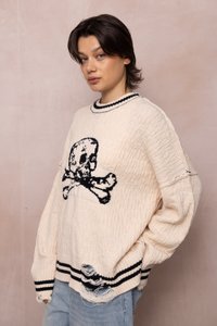 Joven con pelo corto y oscuro, vistiendo un suéter de punto color crema con un diseño de calavera negra y dobladillo desgastado, de pie contra una pared beige lisa.