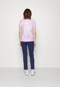 Osoba stojąca tyłem, ubrana w różową bluzkę z krótkim rękawem w stylu tie-dye, granatowe spodnie i białe trampki, na jednolitym tle.