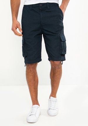 Homme portant un short cargo bleu marine foncé et des baskets blanches, debout devant un fond blanc uni.
