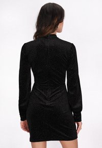 Czarna aksamitna sukienka z długimi rękawami, z teksturowanym wzorem w diamenty i dopasowanym krojem. Wysoki kołnierz i subtelny detal zapięcia z tyłu.