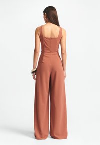 Rostfarbener Overall mit weitem Bein, quadratischem Rückenausschnitt und ärmellosem Design. Hergestellt aus glattem, leichtem Stoff in einer Terrakotta-Farbe.