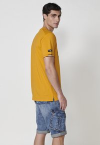 Kreminiai geltonos spalvos trumpomis rankovėmis marškiniai su apvaliu kaklu, papuošti dryžuotais akcentais ant rankovių. Sujungti su šviesiai mėlynomis džinsinėmis šortais su kišenėmis.