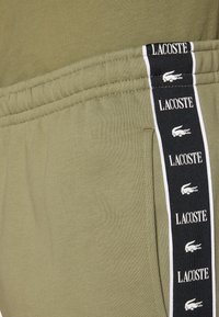 Pantalones de chándal verde oliva con una franja lateral negra que presenta el texto y el logo de "Lacoste" en repetición. Material suave, cintura elástica y bolsillos.