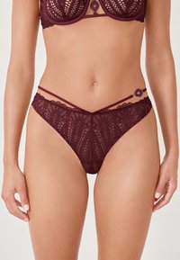 String en dentelle bourgogne avec des motifs floraux complexes, des fines bretelles et un accent en métal circulaire sur le côté. Tissu transparent et texturé.