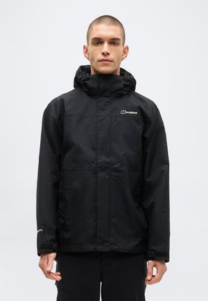 Mlad moški s kratkimi lasmi, ki nosi črno vodoodporno jakno Berghaus s kapuco in stoji pred enobarvnim svetlim ozadjem.