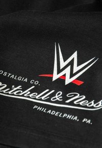 Schwarzer Stoff mit einem hervorgehobenen weißen "WWE"-Logo, roten Akzenten und dem Schriftzug "Mitchell & Ness", mit einer glatten, strukturierten Oberfläche.