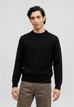 Un homme se tient devant un fond gris clair, vêtu d’un pull en maille noir et d’un pantalon taupe.