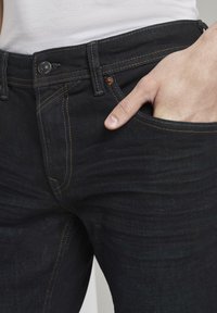 Los pantalones vaqueros de mezclilla oscuros presentan un ajuste slim, costuras naranjas, diseño de cinco bolsillos y un cierre de botón metálico en la cintura.
