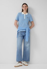 Frau, die gegen eine graue Wand steht und ein blau-weiß gestreiftes Poloshirt, hellblaue Jeans, weiße Sneakers und einen blauen Pullover um die Taille gebunden trägt.