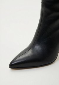 Černá kožená kotníková bota s špičatou špičkou, hladkým povrchem a zakřiveným šitím na bílém pozadí.