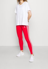 Czerwone legginsy sportowe z trzema białymi paskami po bokach, w połączeniu z luźną białą koszulką. Miękka tkanina, dopasowany krój.