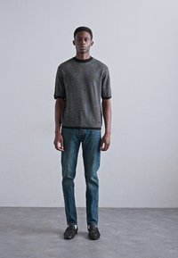 Maglione corto grigio a maniche corte con texture e rifiniture nere, abbinato a un paio di jeans blu e scarpe nere. Il modello è in piedi su uno sfondo semplice.