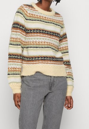 Personne portant un pull tricoté rayé multicolore avec des motifs crème, verts, marrons et noirs, associé à un jean gris.