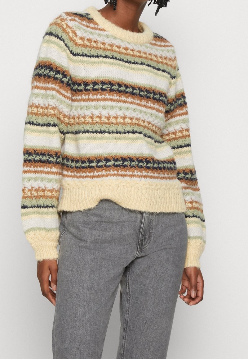Personne portant un pull tricoté rayé multicolore avec des motifs crème, verts, marrons et noirs, associé à un jean gris.