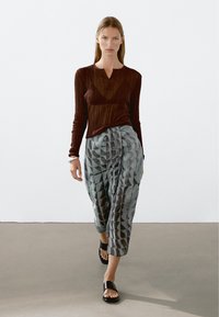 Bruine geribbelde langemouwen top, gecombineerd met grijze patroon wijdere broek met geometrische vormen, en zwarte platte sandalen.
