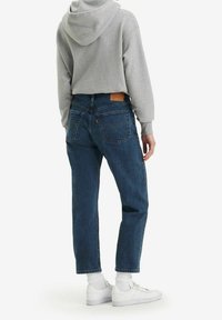 Sweat à capuche gris, jean en denim bleu avec une coupe décontractée, couture visible et patch en cuir de marque à l'arrière, associé à des baskets blanches.