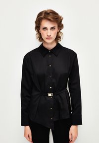 Camicia nera a maniche lunghe con colletto a punta, dotata di bottoni dorati e accentuazione con cintura legata. Tessuto liscio con un design strutturato.
