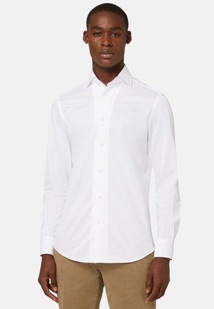 Camisa blanca de manga larga con botones y cuello clásico, hecha de tela suave, con botones claros y un dobladillo curvado. Llevada con pantalones beige.