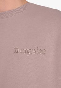 Svetlo roza pulover z rebrastim ovratnikom, na sprednji strani z vezenim napisom "Dropsize" v kontrastni bež gotski pisavi.