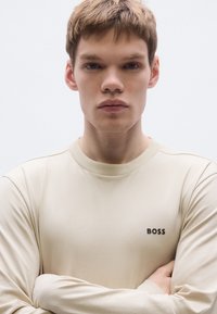 Bézs hosszú ujjú póló, pamut keverékből készült, kerek nyakkivágással és a bal mellrészen fekete "BOSS" logóval.
