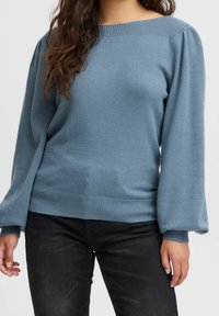 Niebieski sweter z dzianiny z łódkowym dekoltem, ściągaczami na mankietach i dole. Charakteryzuje się bufkami na rękawach i miękką fakturą. Zaprezentowany w połączeniu z czarnymi spodniami.