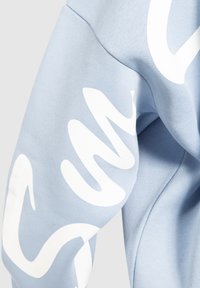 Lichtblauwe sweatshirt met een witte golvende tekstontwerp op de mouw, vervaardigd uit een zachte stof met een gladde textuur en een casual pasvorm.