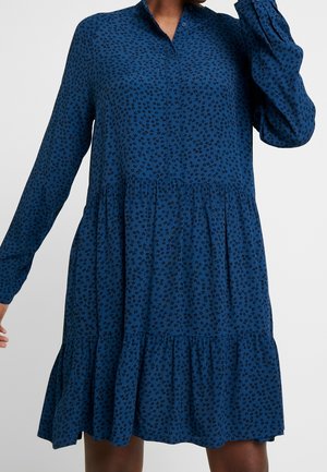 Frau trägt ein langärmliges, blaues Kleid mit Knopfleiste, schwarzem Blumenmuster und gerafften Stufen an Taille und Saum.