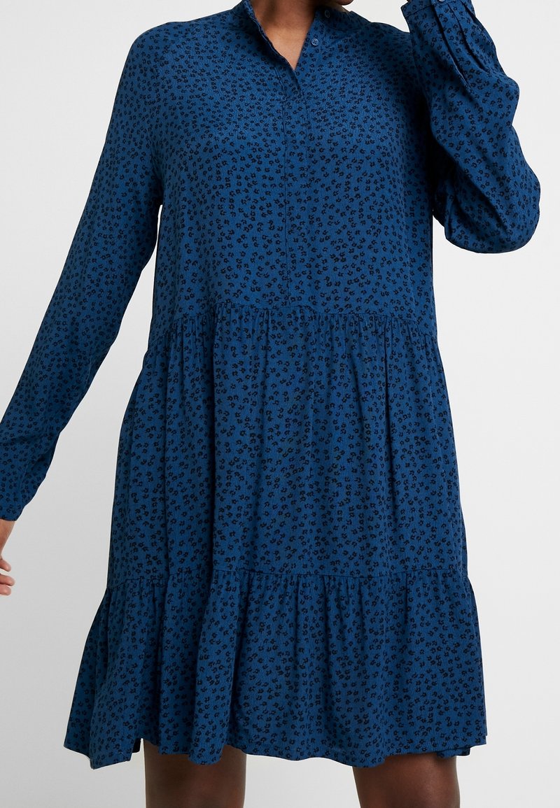 Femme portant une robe bleu à manches longues avec boutons, motif floral noir et volants froncés à la taille et à l’ourlet.
