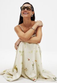 Svetlo béžové kvetinové šaty s plynulou, textúrovanou látkou. Obsahuje zelené a oranžové vzory. Modelka nosí veľké čierne slnečné okuliare a strieborné šperky.