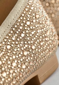 Chaussure beige recouverte densément de strass facettés scintillants, avec des accents en fil d'or et une bordure en tissu texturé.