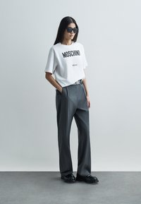 Biała koszulka Moschino z czarnym logo, zestawiona z wysokimi szarymi spodniami i czarnymi butami. Model nosi czarne okulary przeciwsłoneczne.